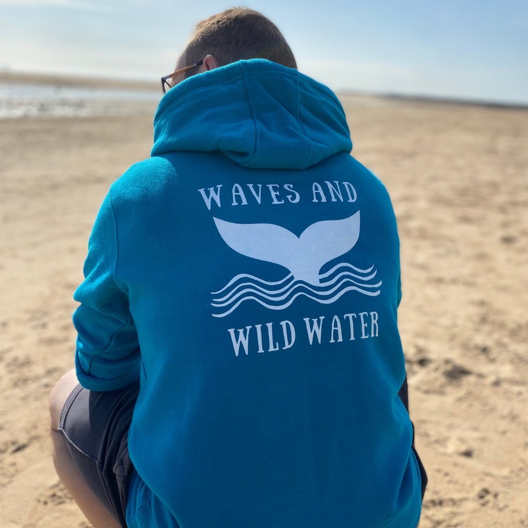 Wild swimmer and Waves and Wild Water owner Glenn, squats on the beach in Wales, whilst wearing his heavyweight hoodie. The super-soft Ocean Depths blue of his hoodie perfectly compliments the white whale tail and waves logo design across the back.