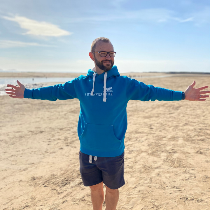 Glenn embraces the welcome sun on the beach in Wales, and stretches his arms out to make the most of the warmth. He wears his Ocean Depths hoodie with white logo print across the chest, and contrasting white inner hood and draw cord. This hoodie is loved by wild swimmers and sea dippers alike.