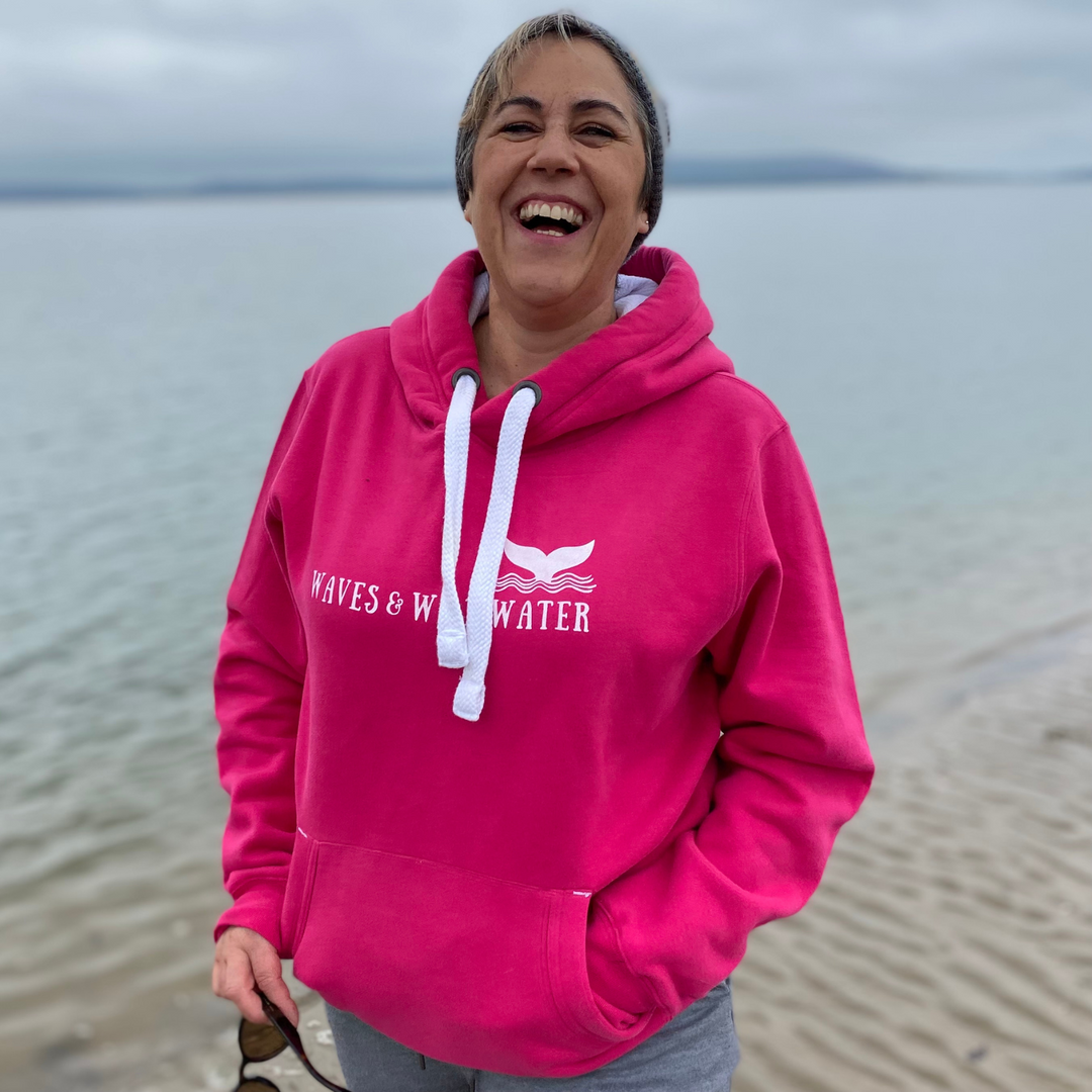 Wild swimmer and open water enthusiast, Claire, wears her Water Lily pink Waves and Wild Water hoodie on the beach. It's a grey day so she's happy she's wearing something so warm and snuggly.