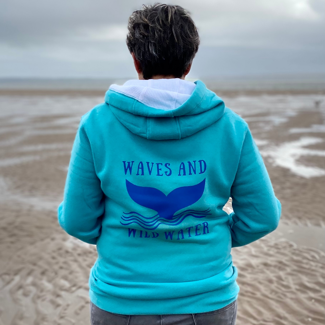 Wild swimmer Claire, looks out over an empty beach, whist wearing her Waves and Wild Water hoodie in lagoon blue. The hoodie has the majestic whale tail logo printed across the back in deep blue water based ink.